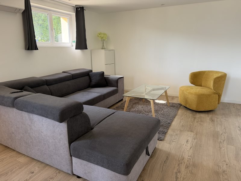 L'espace de consultation : un salon clair, canapé d'angle gris, fauteuil jaune et table basse, sous la lumière naturelle d'une fenêtre.